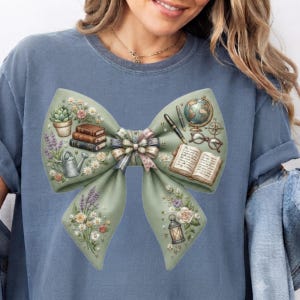 Peut inclure: Un t-shirt bleu avec un grand motif de nœud vert sauge. Le nœud est orné d'illustrations de livres empilés, d'une plante succulente en pot, d'un arrosoir, de lavande, d'un globe, d'un stylo plume, de lunettes, d'un livre ouvert et d'une lanterne.