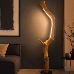 Może przedstawiać: Lampa podłogowa wykonana z naturalnej, jasnej gałęzi drewnianej z pionowym paskiem światła LED wbudowanym wzdłuż jej krzywizny. Gałąź jest zamontowana na okrągłej, czarnej metalowej podstawie, która zapewnia stabilność całej konstrukcji.