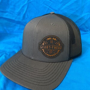 May include: Gray trucker hat with a black mesh back. The front of the hat features a brown leather patch with the words "GEAR HEAD" and a gear design. The hat has a curved brim and a snapback closure.