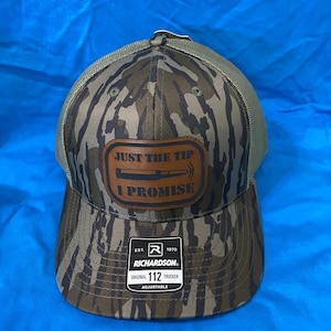 May include: A camouflage trucker hat with a brown leather patch that reads "JUST THE TIP I PROMISE". The hat has a mesh back and a curved brim. The brand "RICHARDSON" is visible on the tag.