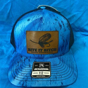 May include: Blue trucker hat with a mesh back and a curved brim. The front panel features a brown leather patch with a fishing lure design and the text "BITE IT BITCH". The hat also has a Richardson patch that says "ORIGINAL 112 TRUCKER".