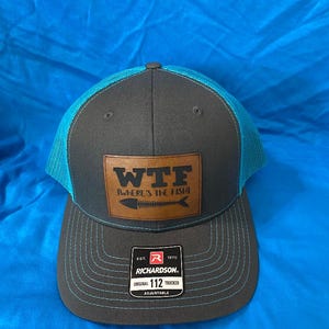 May include: Gray and teal trucker hat with a leather patch that reads "WTF Where's the Fish" with a fish skeleton graphic. The hat has a curved brim and a mesh back. The Richardson brand label is visible.