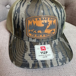 May include: A camouflage print trucker hat with a tan mesh back and a brown leather-like patch on the front. The patch features a silhouette of a deer in a forest with the text IT'S A BAD DAY TO BE A SPIKE. A white sticker displays 112P PRINTED TRUCKER OSFM.