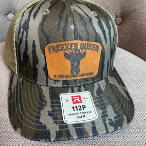May include: A camouflage trucker hat with a tan mesh back and a brown leather patch on the front. The patch features a deer head graphic with a crown and the text "FREEZER QUEEN" and "IF SHE BLOWS SHE GOES". A white product tag is attached to the brim.