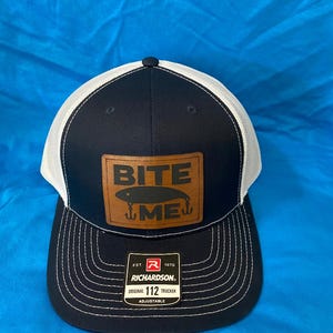 May include: Navy blue and white trucker hat with a brown leather patch. The patch features the words "BITE ME" above a fishing lure graphic. The hat is a Richardson Original 112 Trucker, adjustable.