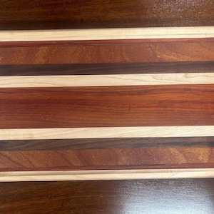 Peut inclure: Planche à découper rectangulaire en bois avec une rainure à jus. Le bois présente des rayures alternées de couleurs marron clair, moyen et foncé. Les bords sont de couleur bois plus claire, posée sur une surface marron foncé.