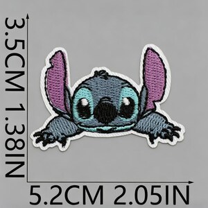K&ouml;nnte beinhalten: Ein gestickter Aufn&auml;her eines blauen Alien-Charakters mit gro&szlig;en violetten Ohren, schwarzen Augen und einer schwarzen Nase. Der Aufn&auml;her misst 3,5 cm mal 5,2 cm, was 1,38 Zoll mal 2,05 Zoll entspricht, wie durch die sichtbaren Ma&szlig;angaben belegt.
