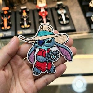以下が含まれることがあります： 青いエイリアンのキャラクターが刺繍されたワッペン。ベージュのつば広帽子にターコイズブルーのバンド、赤いシャツを着用している。キャラクターは両手で小さな灰色のカメラを持っており、紫色の裏地がついた大きな耳が特徴。