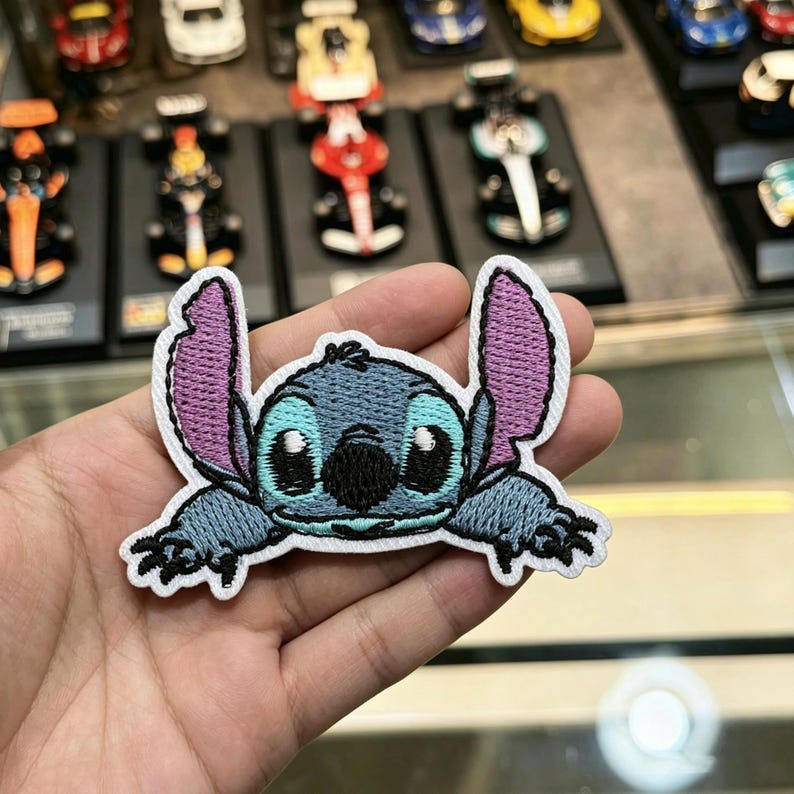 K&ouml;nnte beinhalten: Ein gestickter Aufn&auml;her mit der Figur Stitch, die blaues Fell, gro&szlig;e violett gef&uuml;tterte Ohren, eine schwarze Nase und schwarze Augen hat. Das Design ist mit wei&szlig;em Garn umrandet und zeigt die Figur in Bauchlage mit nach vorne gestreckten Armen.