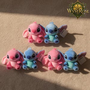 Puede incluir: Seis peluches, tres rosas y tres azules, est&aacute;n dispuestos sobre una superficie marr&oacute;n claro. Los juguetes tienen orejas grandes, ojos grandes y etiquetas peque&ntilde;as. El logotipo de WyseRose es visible en la esquina superior derecha.