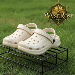 May include: A pair of cream-colored clogs with multiple ventilation holes, resting on a black metal rack. The clogs have a strap for secure wear. The background is green grass. The logo "WyseRose" is visible.
