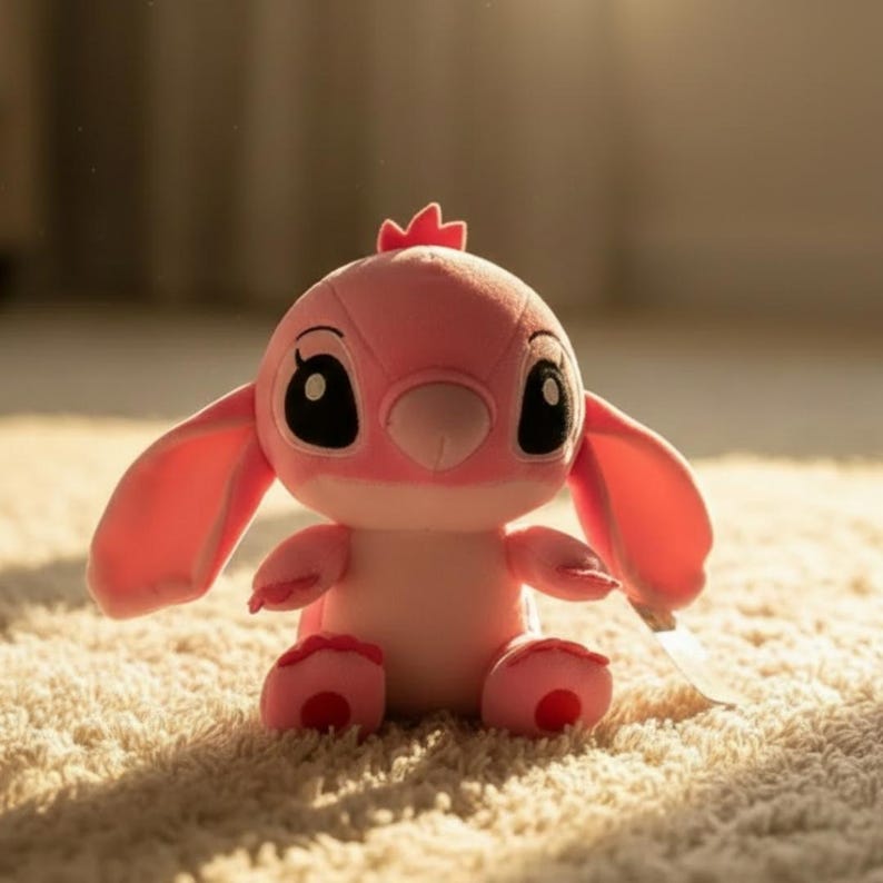 Puede incluir: Un peluche rosa de un personaje de dibujos animados con grandes ojos negros, orejas largas y una peque&ntilde;a corona. El juguete est&aacute; sentado sobre una alfombra color crema. El personaje tiene una nariz rosa y sostiene una peque&ntilde;a etiqueta blanca.