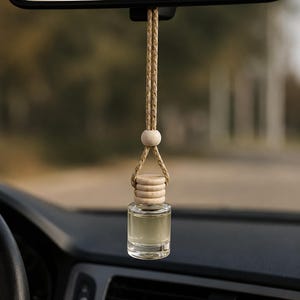 May include: A small, clear glass bottle filled with a light-colored liquid, suspended by a braided tan cord from a rearview mirror. The bottle features a light-colored wooden cap with horizontal ridges and a single round wooden bead on the cord.