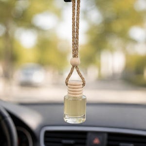Può includere: Una piccola bottiglia in vetro trasparente riempita con un liquido giallo pallido, dotata di un tappo in legno chiaro e una perlina in legno abbinata. La bottiglia pende da un cordino intrecciato color beige con fili dorati, appesa allo specchietto.
