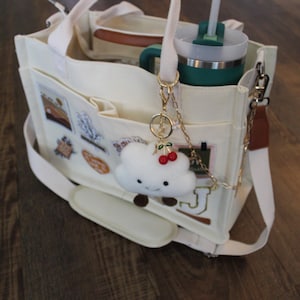 May include: Cream-colored tote bag with multiple pockets and a white shoulder strap. The bag contains a green tumbler with a straw, a lunch container, and a cloud-shaped keychain with cherries. The bag has decorative patches.