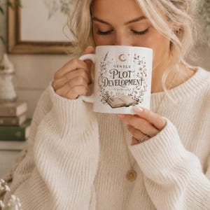 May include: White ceramic mug with the text "Gentle Plot Development" in a decorative font, surrounded by floral and celestial illustrations. The mug features an open book graphic. The mug has a white handle.