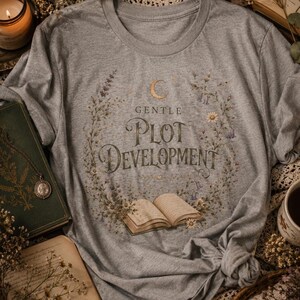 Puede incluir: Una camiseta gris con la frase "GENTLE PLOT DEVELOPMENT" en una fuente decorativa, rodeada por una corona floral y una ilustración de un libro abierto. La camiseta tiene cuello redondo y mangas cortas.