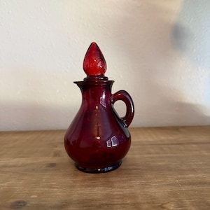 May include: A small, deep red glass cruet with a rounded body, a curved handle on the side, and a matching pointed glass stopper. The glass is translucent and features a smooth, polished finish throughout the entire piece, including the lid.