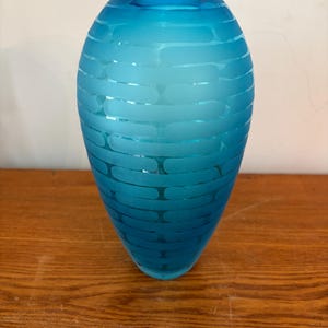 May include: A bright turquoise glass vase with a tapered, rounded silhouette. The exterior surface features a repeating horizontal pattern of frosted, pill-shaped etchings that wrap around the body, creating a textured contrast against the smooth glass.