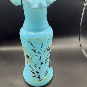 Puede incluir: Un florero de vidrio azul claro con un borde ondulado y fruncido. El cuerpo del florero presenta un diseño floral pintado a mano con hojas de color verde oscuro, tallos negros y una flor pálida. El vidrio tiene un acabado opaco y liso.