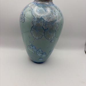 May include: A ceramic vase with a bulbous body and a narrow neck, featuring a light blue glaze decorated with white crystalline patterns that resemble flower-like formations. The rim of the vase has a warm, brownish-orange glazed finish.