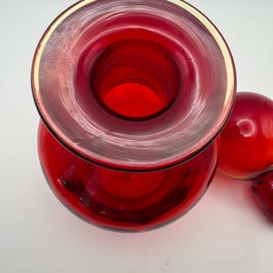 May include: A vibrant red glass decanter with a wide, circular opening and a smooth, polished rim. The decanter features a rounded body and is accompanied by a matching red glass stopper, shown resting beside the main vessel from a top-down view.