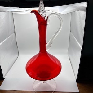 May include: A tall, vibrant red glass decanter featuring a bulbous base, a slender neck, and a clear glass handle. The decanter includes a clear, spiraled glass stopper that sits atop the opening, all supported by a circular clear glass base.