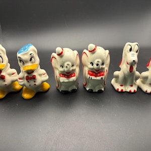 May include: A set of six ceramic salt and pepper shakers featuring cartoon characters. Two white ducks with blue caps, yellow beaks, and red bow ties, two white elephants with red accents, and two white dogs with red ears and tongues are arranged in a row.