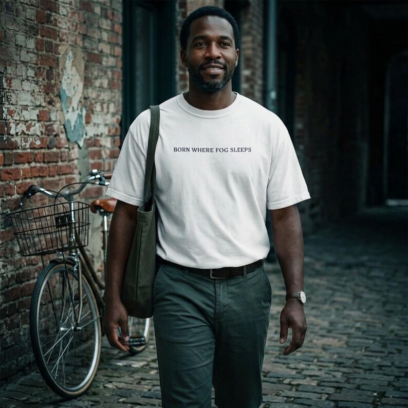 Man walking down a cobblestone alley past a vintage bicycle, wearing a relaxed-fit white t-shirt with "BORN WHERE FOG SLEEPS" in small dark serif lettering. Stardust Frog Hollow Lampkeeper's Journal saying tee.
