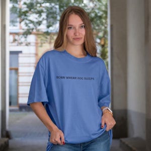 Woman with long hair standing in soft daylight, wearing an oversized cornflower blue t-shirt with "BORN WHERE FOG SLEEPS" in small black serif lettering across the chest. Stardust Frog Hollow Lampkeeper's Journal saying tee.