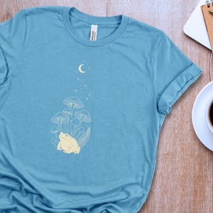 May include: A light blue t-shirt featuring a cream-colored graphic print of a toad sitting beneath a cluster of mushrooms, a fern leaf, and a crescent moon with small stars above the toad. The shirt has a crew neckline and short sleeves.