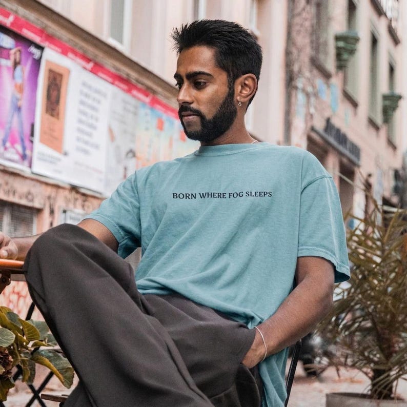 Man seated outdoors in a European street, wearing an oversized dusty teal t-shirt with "BORN WHERE FOG SLEEPS" printed across the chest in small black serif lettering. Stardust Frog Hollow Lampkeeper's Journal saying tee.