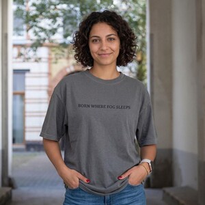 Woman with curly hair smiling in a stone archway, wearing an oversized washed charcoal grey t-shirt with "BORN WHERE FOG SLEEPS" in small dark serif lettering. Stardust Frog Hollow Lampkeeper's Journal saying tee.