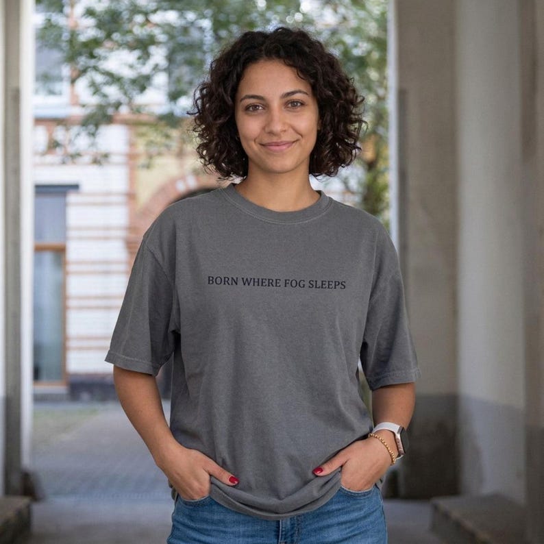 Woman with curly hair smiling in a stone archway, wearing an oversized washed charcoal grey t-shirt with "BORN WHERE FOG SLEEPS" in small dark serif lettering. Stardust Frog Hollow Lampkeeper's Journal saying tee.