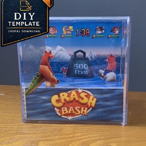 May include: A clear acrylic cube display featuring a Crash Bash video game diorama. Inside, a 3D scene shows characters, a blue ocean, icebergs, and a weight labeled 500 lbs. The front panel displays the Crash Bash logo in yellow and orange text.