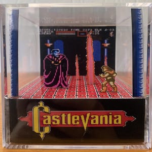 May include: A Castlevania diorama in a clear cube. The scene features pixelated characters and a background from the video game. The Castlevania logo is displayed at the bottom. The cube is on a wooden surface.