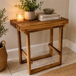 Peut inclure: Une table d'appoint rectangulaire en bois avec un plateau à lattes et un cadre robuste. Sur la table se trouvent une bougie allumée dans un photophore en verre strié, une plante verte en pot, un petit bol en céramique et deux livres empilés.