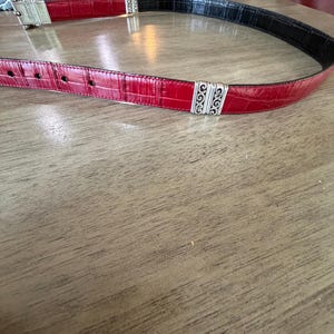 K&ouml;nnte beinhalten: Ein roter und schwarzer G&uuml;rtel mit silberfarbenen Zierakzenten. Der G&uuml;rtel hat ein strukturiertes, krokodilartiges Muster. Die Schnalle ist silberfarben und hat ein dekoratives Design. Der G&uuml;rtel liegt auf einer Holzoberfl&auml;che.