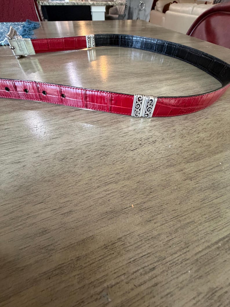K&ouml;nnte beinhalten: Ein roter und schwarzer G&uuml;rtel mit silberfarbenen Zierakzenten. Der G&uuml;rtel hat ein strukturiertes, krokodilartiges Muster. Die Schnalle ist silberfarben und hat ein dekoratives Design. Der G&uuml;rtel liegt auf einer Holzoberfl&auml;che.