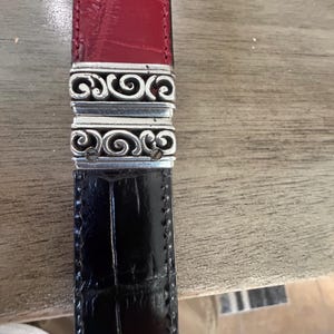 K&ouml;nnte beinhalten: Ein zweifarbiges G&uuml;rtel mit einem roten Oberteil und einem schwarzen Unterteil. Der G&uuml;rtel ist durch einen silberfarbenen Metallverschluss mit einem verzierten, wirbelnden Design verbunden. Das Leder scheint ein strukturiertes, gepr&auml;gtes Muster zu haben.