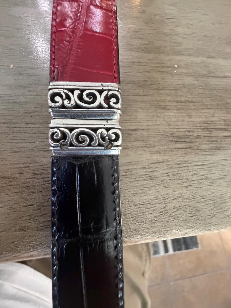 K&ouml;nnte beinhalten: Ein zweifarbiges G&uuml;rtel mit einem roten Oberteil und einem schwarzen Unterteil. Der G&uuml;rtel ist durch einen silberfarbenen Metallverschluss mit einem verzierten, wirbelnden Design verbunden. Das Leder scheint ein strukturiertes, gepr&auml;gtes Muster zu haben.
