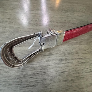 K&ouml;nnte beinhalten: Ein roter Lederg&uuml;rtel mit einer silbernen Schnalle. Die Schnalle hat ein verziertes Design und einen rechteckigen silbernen Verschluss. Der G&uuml;rtel liegt auf einer Holzoberfl&auml;che.