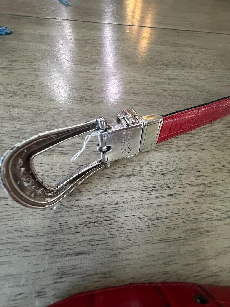 K&ouml;nnte beinhalten: Ein roter Lederg&uuml;rtel mit einer silbernen Schnalle. Die Schnalle hat ein verziertes Design und einen rechteckigen silbernen Verschluss. Der G&uuml;rtel liegt auf einer Holzoberfl&auml;che.