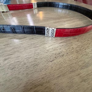 K&ouml;nnte beinhalten: Ein zweifarbiger G&uuml;rtel mit einem schwarz-roten Krokodil-Textur-Design. Der G&uuml;rtel ist mit silberfarbenen Metallbeschl&auml;gen versehen. Der G&uuml;rtel ist etwa 3,8 cm breit.