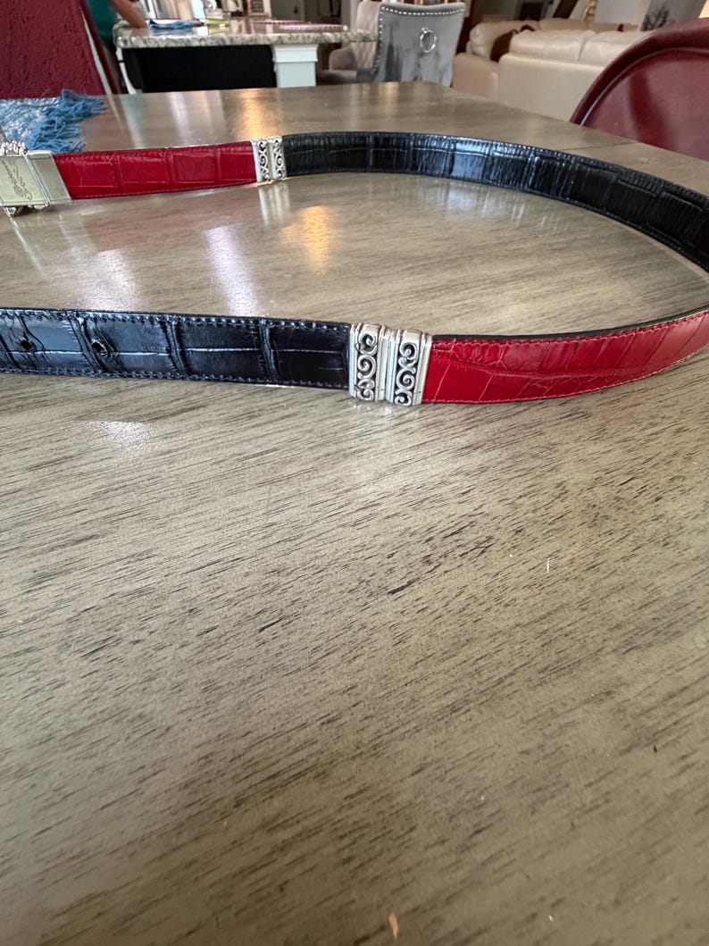 K&ouml;nnte beinhalten: Ein zweifarbiger G&uuml;rtel mit einem schwarz-roten Krokodil-Textur-Design. Der G&uuml;rtel ist mit silberfarbenen Metallbeschl&auml;gen versehen. Der G&uuml;rtel ist etwa 3,8 cm breit.