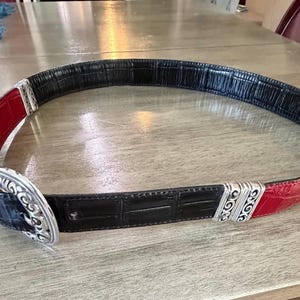K&ouml;nnte beinhalten: Ein schwarzer und roter G&uuml;rtel mit einer silbernen Schnalle. Der G&uuml;rtel hat ein strukturiertes, krokodilartiges Muster. Die Schnalle ist silberfarben mit einem verzierten Design. Der G&uuml;rtel ist etwa 3,8 cm breit.