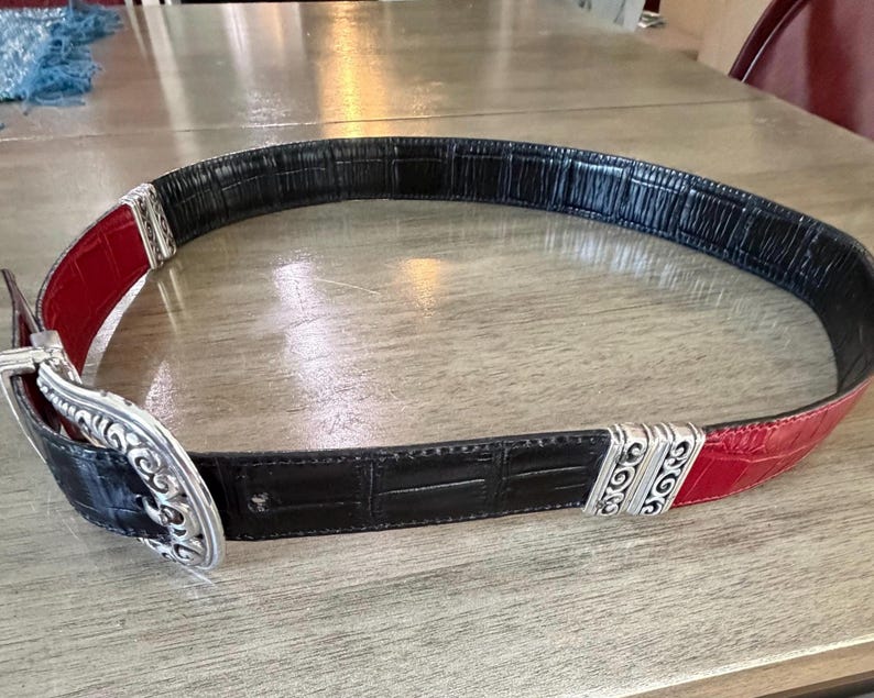 K&ouml;nnte beinhalten: Ein schwarzer und roter G&uuml;rtel mit einer silbernen Schnalle. Der G&uuml;rtel hat ein strukturiertes, krokodilartiges Muster. Die Schnalle ist silberfarben mit einem verzierten Design. Der G&uuml;rtel ist etwa 3,8 cm breit.
