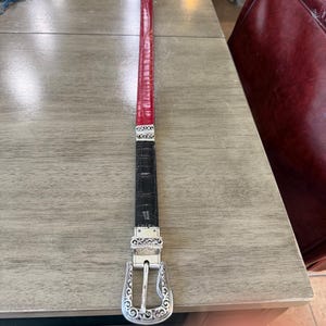 K&ouml;nnte beinhalten: Ein Wendeg&uuml;rtel mit einem roten und schwarzen Lederriemen. Der rote Abschnitt hat ein strukturiertes Muster. Der G&uuml;rtel hat eine silberfarbene Schnalle mit einem verzierten Design. Der G&uuml;rtel liegt auf einer hellen Holzoberfl&auml;che.