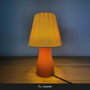May include: A table lamp with a vibrant orange, textured, honeycomb-patterned shade and a matching hourglass-shaped base. The lamp emits a warm, glowing light, highlighting the intricate geometric surface detail across both the shade and the base.