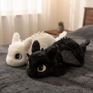 May include: Two plush dragon toys, one white and one black, rest on a gray textured surface. The white dragon has large, round eyes with blue accents. The black dragon has yellow eyes and small wings. Both have soft, plush bodies.